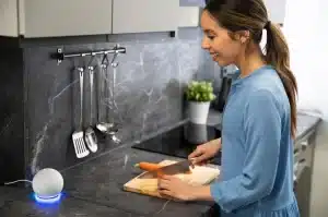 Mujer usando un asistente de voz inteligente mientras cocina, ejemplo de tecnología práctica en el hogar.