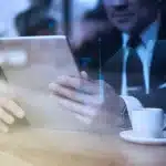 Hombre usando una tablet en un ambiente de oficina con gráficos digitales y tecnología avanzada, acompañado de una taza de café. Aprovechando las ventajas de la tecnología en su negocio.