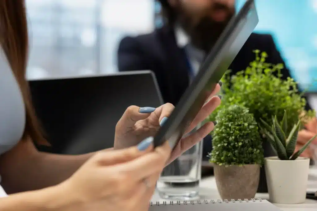 Mujer usando una tablet en una mesa de oficina, con plantas y elementos de trabajo alrededor, mientras una persona está parcialmente visible al fondo. Aprovecha las ventajas de la tecnología