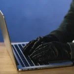 Individuo con guantes escribiendo en un portátil representando un ciberdelincuente realizando una estafa en internet