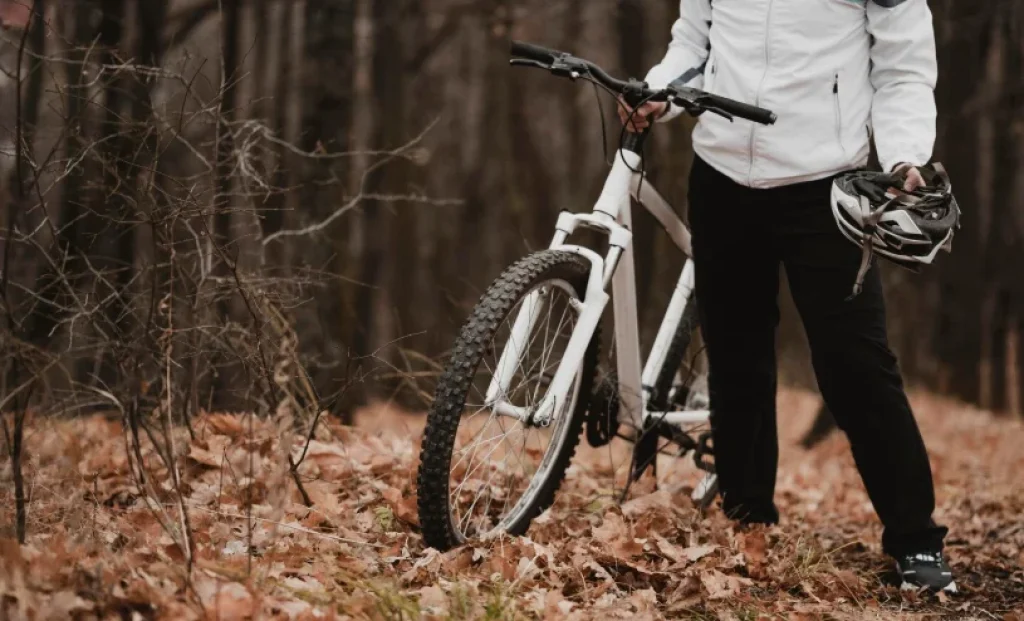 Persona caminando con una bicicleta de montaña por un bosque con hojas secas, llevando casco en la mano, actividad física y ciclismo cotidiano al aire libre